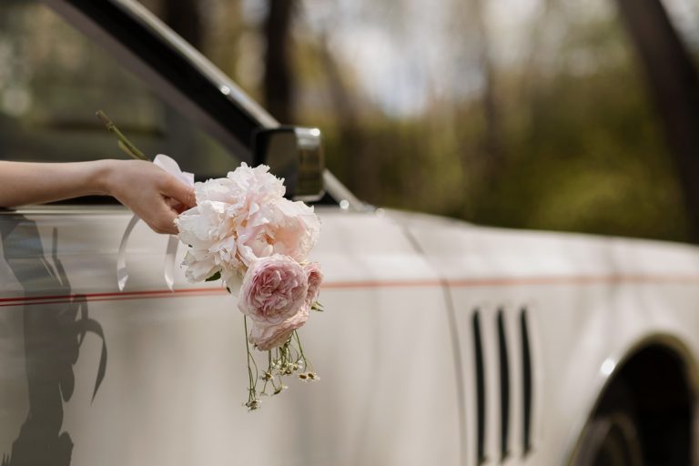 Hand of bride holding bouquet sticking out of a car window illustrates blog "Does Being Married Impact Your Auto Insurance Rates?"